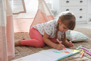Little girl drawing in sketchbook near toy wigwam at home © New Africa