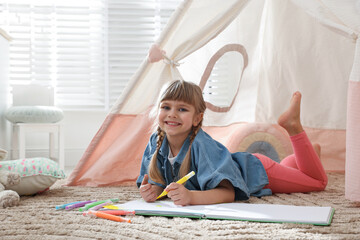 Little girl drawing in sketchbook near toy wigwam at home © New Africa