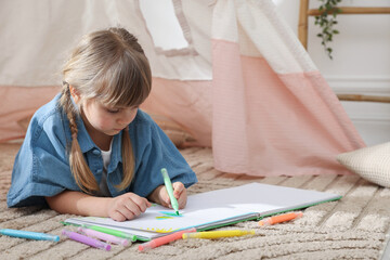 Little girl drawing in sketchbook near toy wigwam at home © New Africa