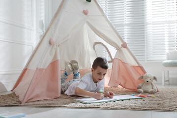Little boy drawing in sketchbook near toy wigwam at home © New Africa