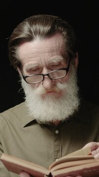 Mature Caucasian man wearing glasses. Flipping pages of old book. Male holding book and reading carefully. Looking down with focused expression. Standing in dark background in studio.