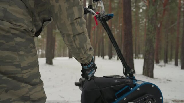 Caucasian rider standing on tracked atv, snowcovered pine forest, camouflage jacket and sturdy boots, inspecting tracks and stance, preparing for extreme offroad ride, muted cold light and quiet