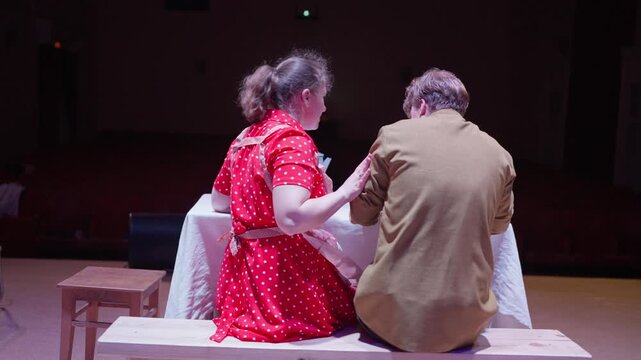 Emotional stage performance of two actors embracing at a table, expressing intimacy and vulnerability under dramatic theater lighting during a heartfelt scene