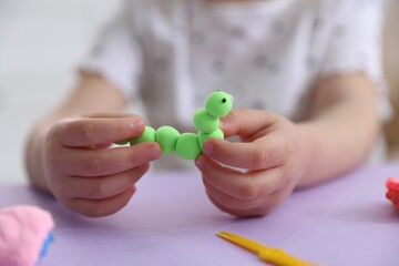 Little girl with caterpillar made of play dough at violet table indoors, closeup