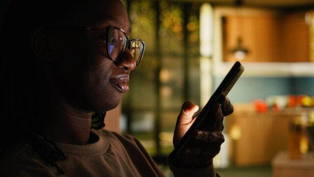 African American young woman relaxing alone and checking apps, scrolling social media on phone at home. Comfortable domestic life highlighting calm cozy lifestyle, modern everyday tranquility.