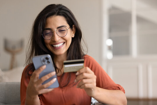 Happy Latina woman sits in living room with card and smartphone, making online purchase, paying bill through banking app, managing subscription services, sending money via mobile payment application