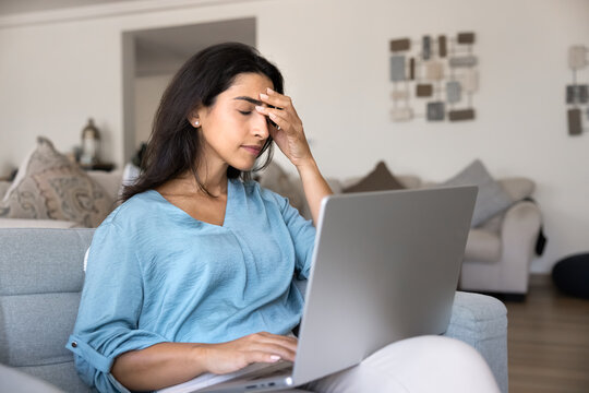 Woman sits on sofa at home, experiencing stress, overwhelmed by tasks or remote work pressure, frustrated with slow or malfunctioning laptop, got distressing financial information, disappointing news