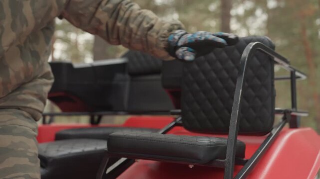 Camo rider climbing onto red atv, gloved hand gripping metal rail, quilted seat and wet fender, pine forest backdrop, overcast drizzle, rugged gear preparing for offtrail adventure