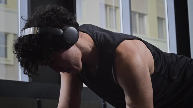 Close up of young Asian man listening to music while doing one arm dumbbell row on bench in modern gym