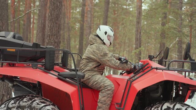 Atv rider in snowy pine forest revs throttle, camo jacket and white helmet visible, red quad with chunky tires stationary between trees, closeup of hands on handlebar, technical focus and adrenaline