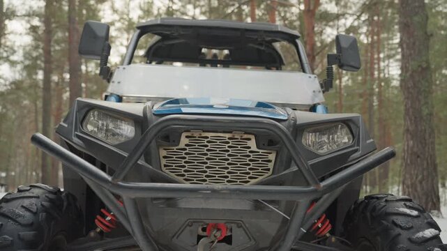 Utv front grille closeup in forest, bumper, winch and headlights framed by snow and pine, crew inspects suspension and engine while driver adjusts helmet, metal textures and rugged tires emphasized,