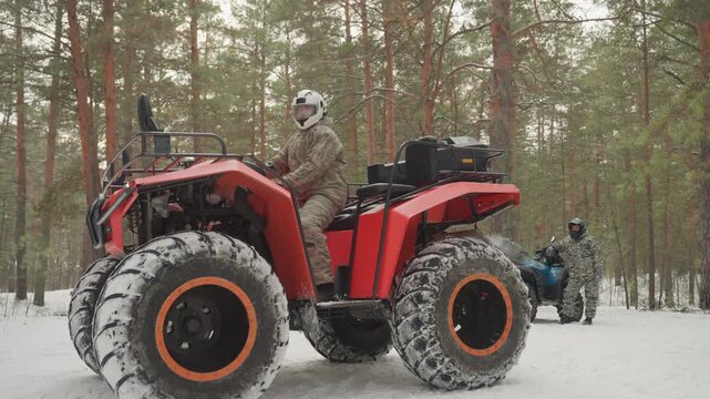 Red atv in snowy pine forest, parked with helmeted riders nearby, oversized studded tires rimmed with snow, cold winter light filtering through tall pines, rider performing pre ride check, rugged