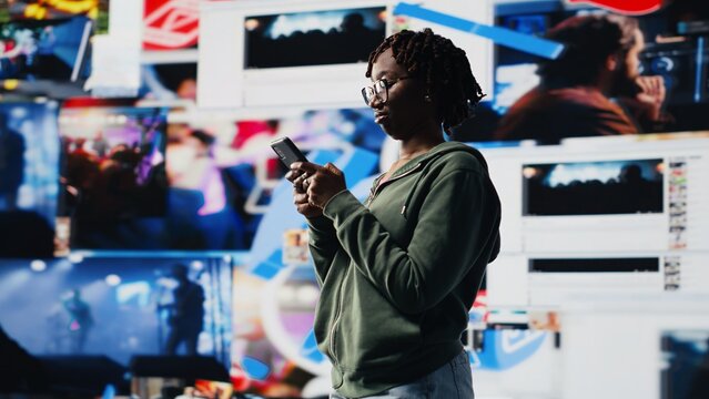 Happy woman uses smartphone to do texting with friends, discussing movies, standing in front of vibrant video wall collage. Teenager chatting with other social media users using mobile phone, camera B