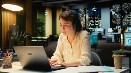 Fototapeta premium Businesswoman taking headphones out and stretching her back at desk, correcting her posture after working on tasks with ambience music. Woman with muscle stiffness and tension. Camera A.