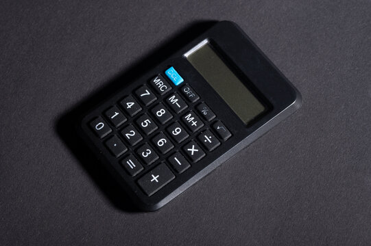 A black calculator rests on a dark surface. It features a standard layout with buttons for numbers and basic operations. The clear screen is ready to display results from calculations. This tool is co