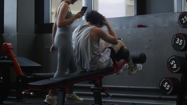 Medium shot of young Asian man having bench abs workout while his girlfriend fooling around and recording video of their training in gym