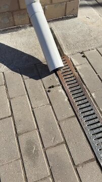 White pipe metal grate on pavement, patterned metal grate covers spillway installed across paving stones on footpath, sunlight.