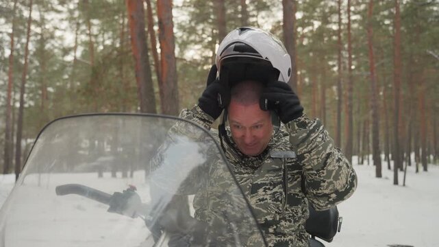 Caucasian man removing helmet next to atv in snowy pine forest, camouflage jacket, adjusting visor and gloves, smiling and breathing visible, relaxed mood after short ride, closeup portrait showing