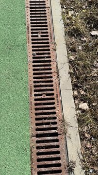 Closeup sports field drainage grate showing rusted iron slots, debris lodged along turf edge, concrete border, textured artificial.