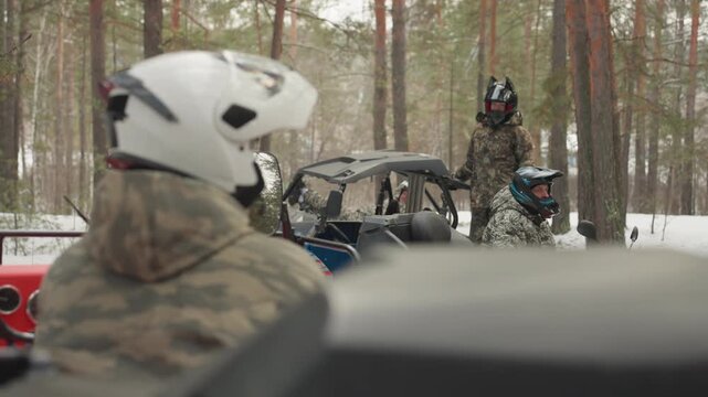 Red atv driver follows convoy through snowy pine trail, helmeted rider grips handlebars while watching blue utv and camo crew, engines rumble and throttle hum, cold wind and overcast sky heighten