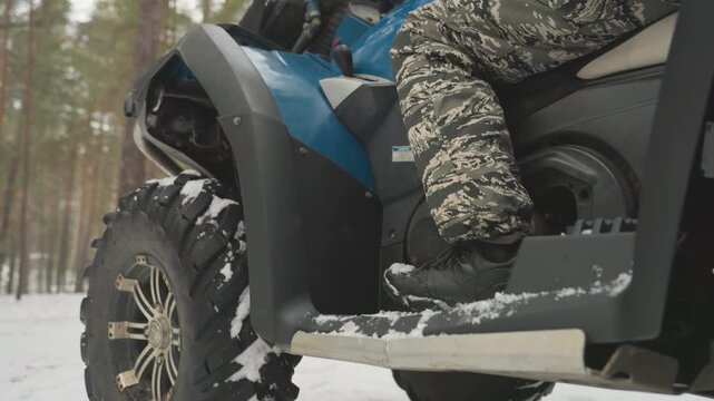Blue atv rider in snowy forest closeup of quad footrest and snow covered tires, camo pants on peg, lowangle rear shots showing rugged tread and suspension, pine trees in background, crisp cold light