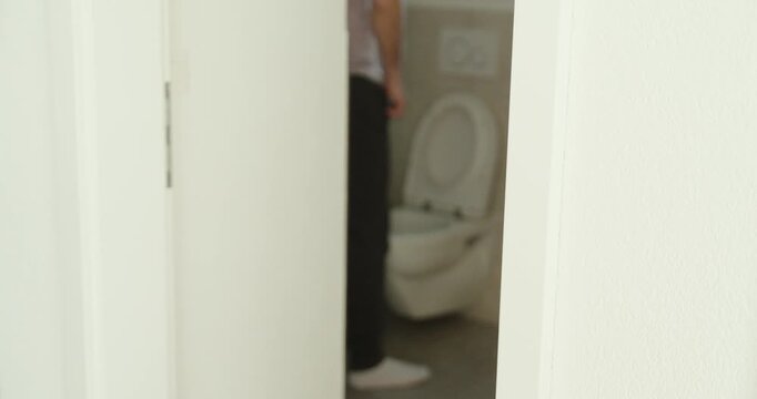 View through a slightly open door of a man standing and using a toilet with the seat lifted, representing everyday bathroom routine, hygiene behavior, and privacy.