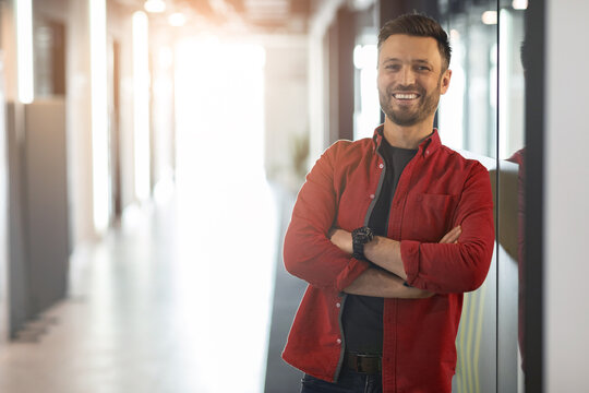 A young man stands with arms crossed in an office hallway. He wears a red shirt and looks directly at the camera with a smile. The hallway has bright lighting and modern features.