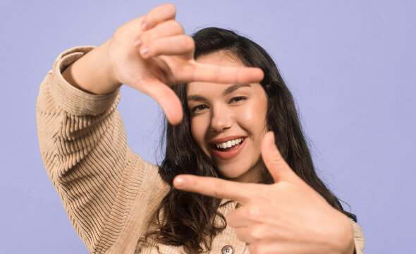 A young woman smiles as she frames her face with her hands. She stands in a studio with a light purple backdrop. Her expression shows joy and playfulness during a fun moment.