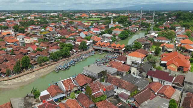 Aerial view of boats docked along the river contrasting with the red-tiled rooftops of buildings and the green trees, Jepara, Central Java, Indonesia.