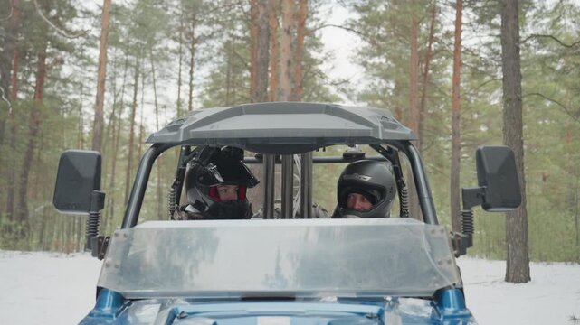 Front view atv cockpit helmets, two riders in protective gear sit inside utv windshield in snowcovered pine forest, driver glances at passenger while passenger adjusts visor, tense anticipation