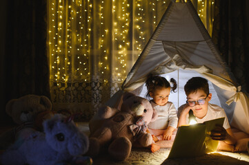 Siblings reading book in teepee at night © Serhii