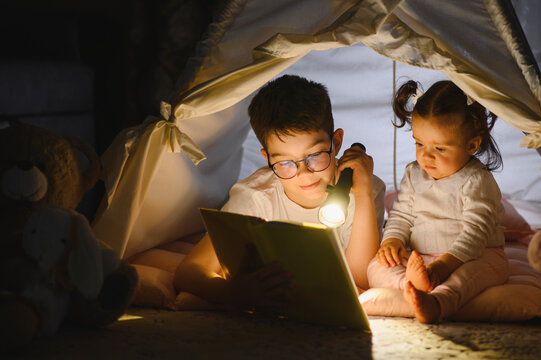 Children reading book with flashlight in blanket fort