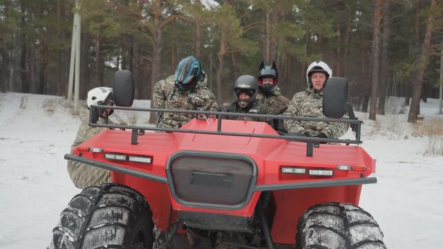 Four riders seated on red quad in snowy forest clearing. Friends in helmets and camo jackets share laughs, ready for offroad adventure, big tires and rugged suspension visible, upbeat camaraderie