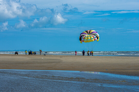 Cox's Bazar, Bangladesh - 18 August 2023: Aerial view of a colorful parasail drifting gently above the sandy beach, as people gather at the shoreline, where the blue sky meets the ocean.