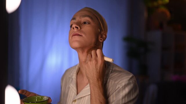 Young man wearing a wig cap applying foundation with a brush. Meticulous preparation in a dressing room before a show or performance