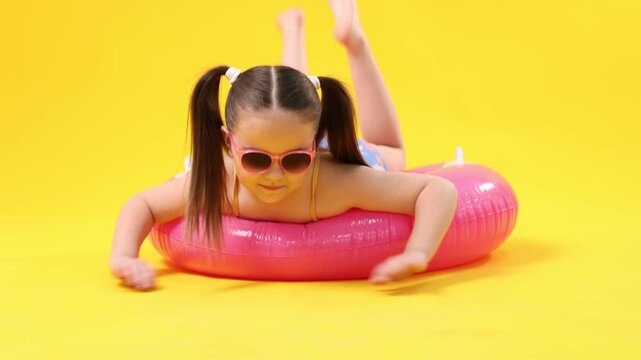 Little girl in swimsuit with inflatable ring and sunglasses on yellow background