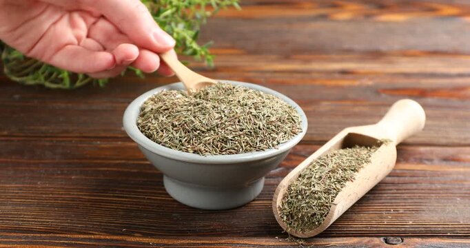 Woman taking dried thyme with spoon from bowl at wooden table, closeup