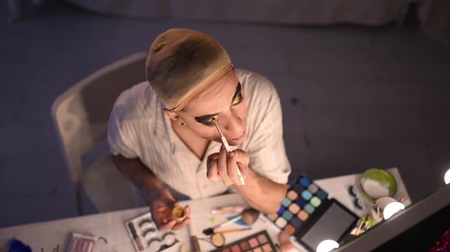 Overhead shot of a youthful man putting on bold makeup as he prepares in a backstage area for his drag show act