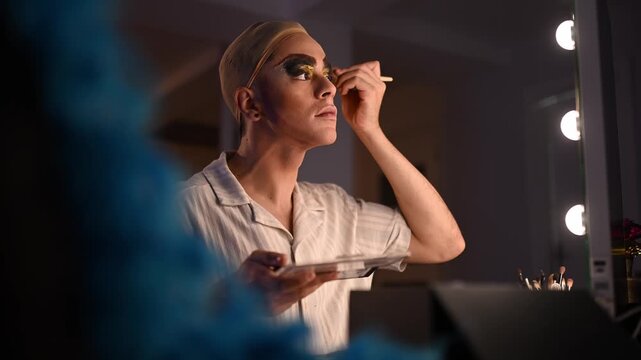 Professional drag queen applying extravagant eyeshadow makeup in a backstage dressing room mirror before the show