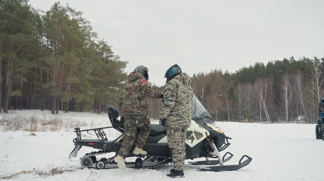 Two white men by snowmobile exchanging handshake and words, snowy open field near pine forest, full winter gear and helmets, one mounting vehicle while partner offers support and checks engine,
