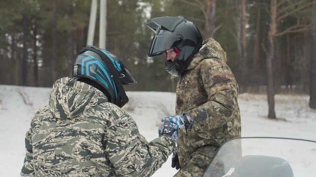 Two riders exchanging words near atv, camouflage gear and helmets on, eye contact and guidance, driver offers short instruction, passenger nods in agreement, windshield frost and pine backdrop, subtle