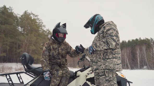 Two riders handshake near snowmobile, camouflage suits and mirrored goggles, conversation about route and safety checks, technician silhouette in background by tree line, atmosphere of mutual trust