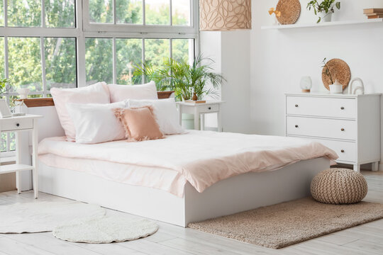 Bed with stylish pink bedding, commode, bedside tables and houseplants near window in bedroom