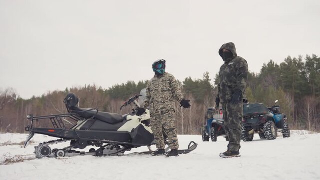 Snowmobile beside atv riders briefing on frozen field, camouflaged participants checking route and gear, distant tree line, overcast light, focused mood and safety checks before offroad run