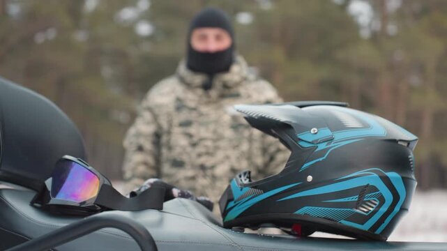 Rider prepares for adventure. Helmet ready before daring journey. Approaching rider gets ready for extreme ride. Camouflage clad rider anticipates intense ATV adventure in winter forest