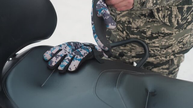 Atv glove on snowmobile seat, camo rider securing strap before extreme ride. Closeup view shows patterned insulated glove rested on padded seat near handlebar, stitched palm, quickrelease clip,
