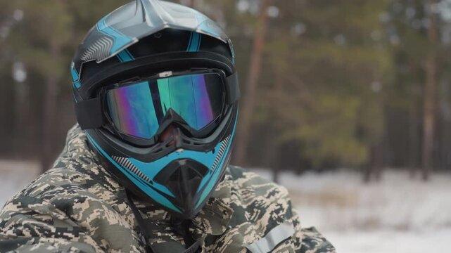 Helmeted rider in snowy pine background stares through mirrored goggles, teal helmet and camouflage jacket, breath visible in cold air, focused expression, quiet preride pause, reflection of trees