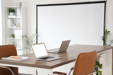 Fototapeta premium Interior of modern conference hall with laptops on table, houseplants and projector screen in light office