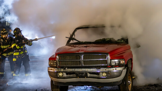 Firefighters work to put out flames coming from a red truck. Smoke fills the air as they spray water on the vehicle. This scene takes place at night in a city setting.