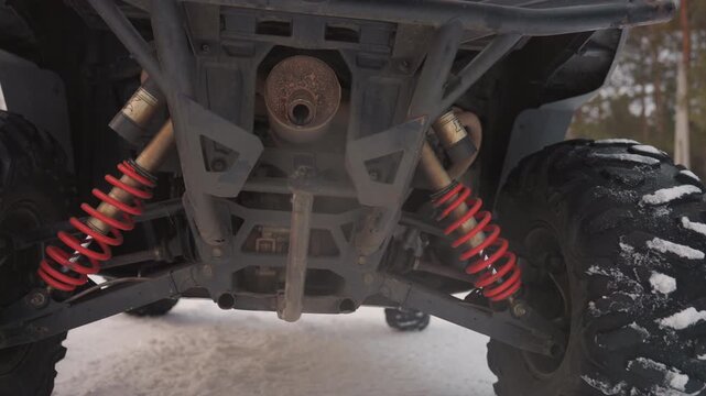 Underside ATV suspension in snow, showing red coil shocks, rugged tread tires, rusted exhaust and bouncing axle on frozen forest trail. Lowangle closeup emphasizes vibration, impact and offroad